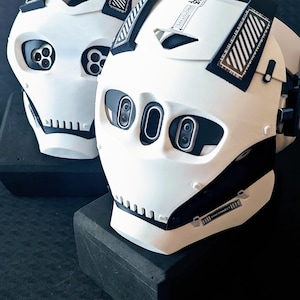 May include: Two white and black full-face masks with a futuristic design. Each mask features three circular lens-like openings in the eye area, a jagged mouth section, and black geometric accents. One mask displays the text PROTOTYPE and DISCONNECT.