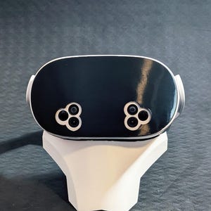 May include: A black and white VR headset with a white base. The headset has a glossy black front with two sets of three circular lenses. The base is white and angular. The overall design is modern and sleek.