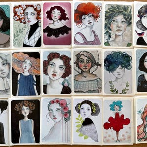 May include: A collection of twenty watercolor portraits of women, each on a rectangular card. The portraits feature various hairstyles and expressions, with a soft color palette of blues, reds, and greens. The artwork has a whimsical and artistic style.