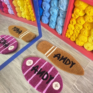 Op de afbeelding: Twee ovale deurmatten met het woord "ANDY" in zwarte letters. De ene deurmat is bruin met witte strepen en de andere is paars met witte strepen. Beide deurmatten hebben een witte ster in elke hoek.