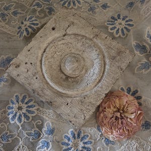 May include: A square, weathered off-white wooden architectural block featuring a central circular medallion with a raised dome. Beside the block sits a single, dried, multi-petaled pale pink rose blossom, all resting upon a piece of sheer white lace.