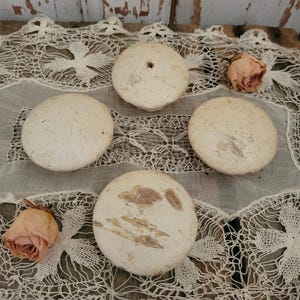 May include: Four off-white, round, wooden objects with a distressed finish are arranged on a white lace doily. Two dried, pink roses are also present. One wooden object has a hole in the center.