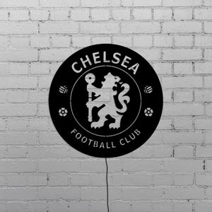 Puede incluir: Un letrero circular negro con el logotipo del Chelsea Football Club, que incluye un le&oacute;n sosteniendo un bast&oacute;n, rodeado por las palabras "CHELSEA FOOTBALL CLUB". El letrero est&aacute; montado en una pared de ladrillos blancos.