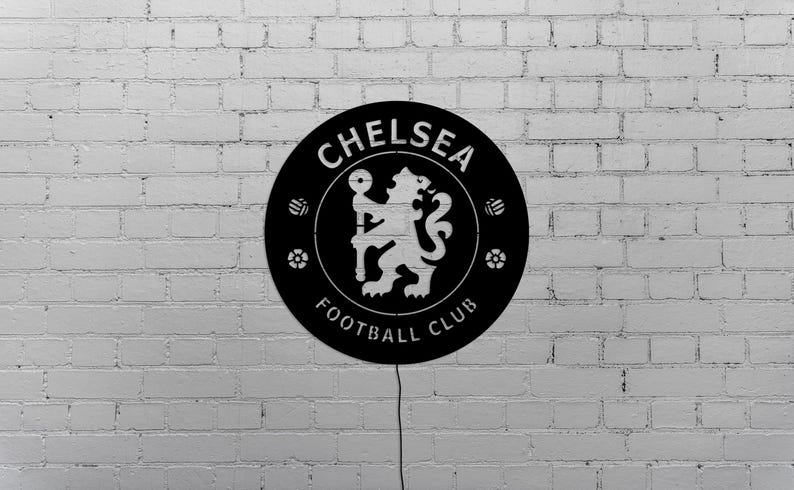 Puede incluir: Un letrero circular negro con el logotipo del Chelsea Football Club, que incluye un le&oacute;n sosteniendo un bast&oacute;n, rodeado por las palabras "CHELSEA FOOTBALL CLUB". El letrero est&aacute; montado en una pared de ladrillos blancos.