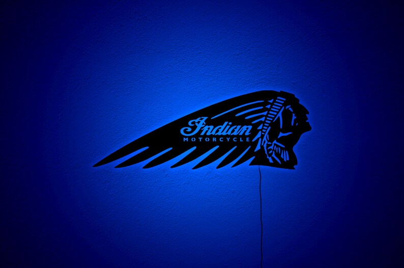 May include: Black metal wall art featuring the Indian Motorcycle logo, illuminated with blue LED lights. The design includes the iconic headdress and wing, with the words "Indian MOTORCYCLE" in a stylized font. The art is mounted on a blue wall.