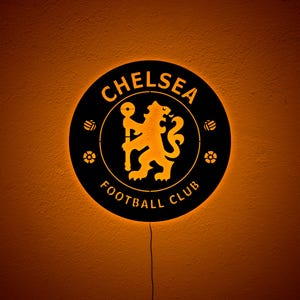 Puede incluir: Un letrero redondo negro del Chelsea Football Club con el logotipo y el texto del equipo en dorado. El letrero est&aacute; iluminado con un brillo c&aacute;lido anaranjado y tiene un cable de alimentaci&oacute;n que se extiende hacia abajo.