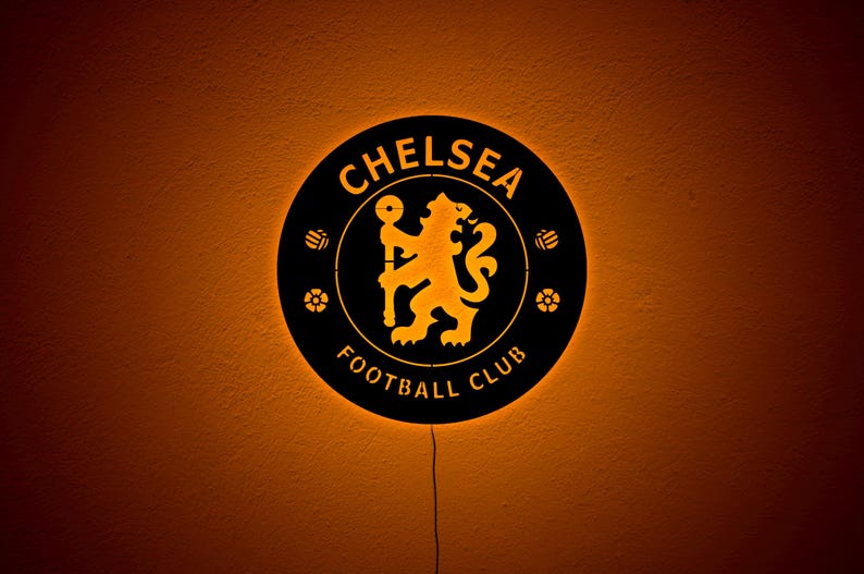 Puede incluir: Un letrero redondo negro del Chelsea Football Club con el logotipo y el texto del equipo en dorado. El letrero est&aacute; iluminado con un brillo c&aacute;lido anaranjado y tiene un cable de alimentaci&oacute;n que se extiende hacia abajo.