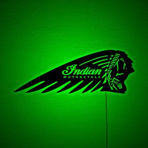 May include: Black metal Indian Motorcycle logo with green neon lighting. The logo features a stylized Native American headdress and the words "Indian MOTORCYCLE" in a script font. The sign is mounted on a green wall.