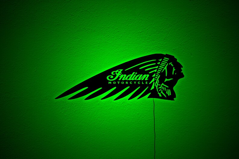 May include: Black metal Indian Motorcycle logo with green neon lighting. The logo features a stylized Native American headdress and the words "Indian MOTORCYCLE" in a script font. The sign is mounted on a green wall.
