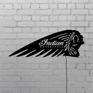 May include: Black metal sign featuring the Indian Motorcycle logo. The sign is shaped like a wing with the words "Indian MOTORCYCLE" and a profile of a Native American chief. The sign is mounted on a white brick wall.