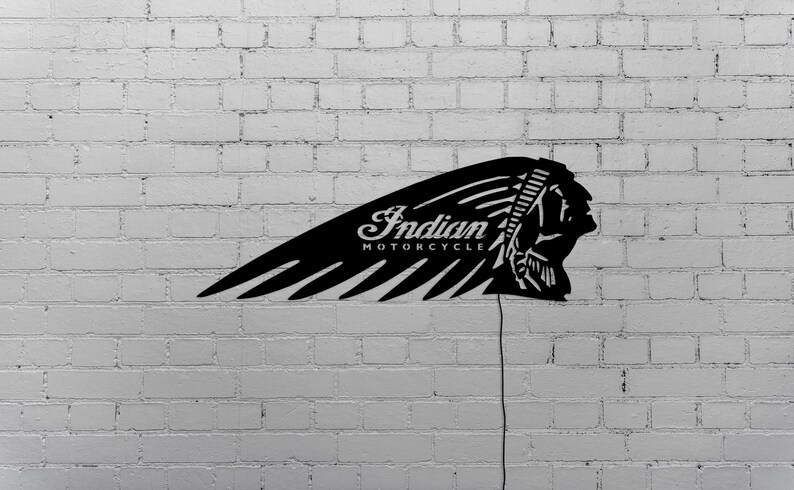 May include: Black metal sign featuring the Indian Motorcycle logo. The sign is shaped like a wing with the words "Indian MOTORCYCLE" and a profile of a Native American chief. The sign is mounted on a white brick wall.