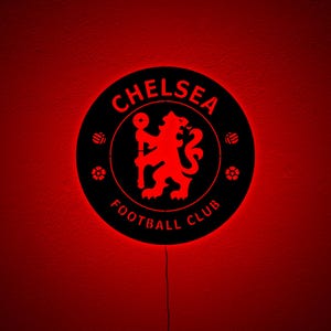 Puede incluir: Un letrero redondo negro del Chelsea Football Club con un logotipo y texto rojo iluminado. El letrero presenta el emblema del club, un le&oacute;n, y las palabras "Chelsea Football Club". El fondo es rojo.