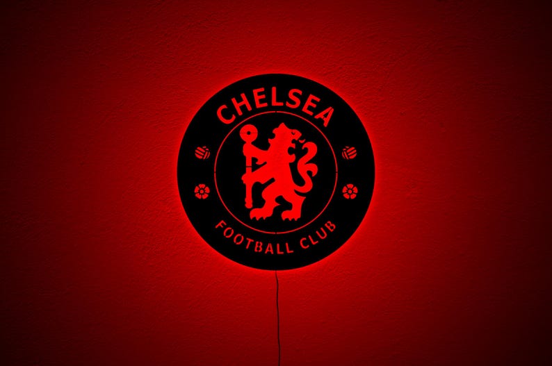Puede incluir: Un letrero redondo negro del Chelsea Football Club con un logotipo y texto rojo iluminado. El letrero presenta el emblema del club, un le&oacute;n, y las palabras "Chelsea Football Club". El fondo es rojo.