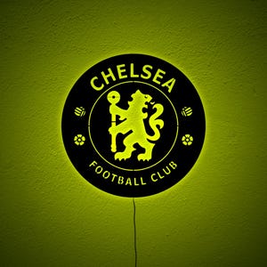 Puede incluir: Un letrero redondo negro del Chelsea Football Club con un logotipo y texto iluminados en amarillo. El logotipo presenta un le&oacute;n sosteniendo un bast&oacute;n. El letrero est&aacute; montado en una pared verde lima texturizada.