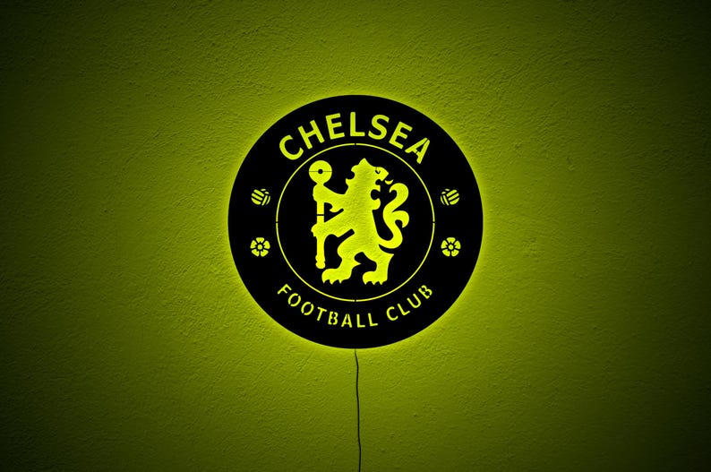 Puede incluir: Un letrero redondo negro del Chelsea Football Club con un logotipo y texto iluminados en amarillo. El logotipo presenta un le&oacute;n sosteniendo un bast&oacute;n. El letrero est&aacute; montado en una pared verde lima texturizada.