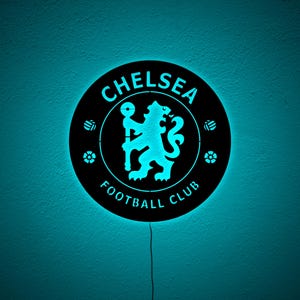 Puede incluir: Un letrero redondo y negro del Chelsea Football Club con un logotipo y texto azul brillante. El letrero presenta el emblema del equipo, un le&oacute;n sosteniendo un bast&oacute;n, y las palabras "Chelsea Football Club". El letrero est&aacute; iluminado con luz azul.