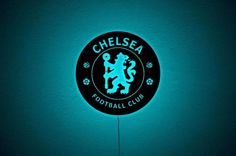 Puede incluir: Un letrero redondo y negro del Chelsea Football Club con un logotipo y texto azul brillante. El letrero presenta el emblema del equipo, un le&oacute;n sosteniendo un bast&oacute;n, y las palabras "Chelsea Football Club". El letrero est&aacute; iluminado con luz azul.