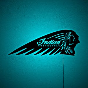 May include: Black Indian Motorcycle logo wall art with a turquoise neon glow. The logo features the text "Indian MOTORCYCLE" in a stylized font, along with the profile of a Native American chief. The wall art is mounted on a turquoise wall.