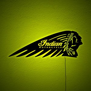 May include: Black Indian Motorcycle logo sign with a yellow glow against a textured green wall. The sign features the brand name and a Native American profile within a wing design.