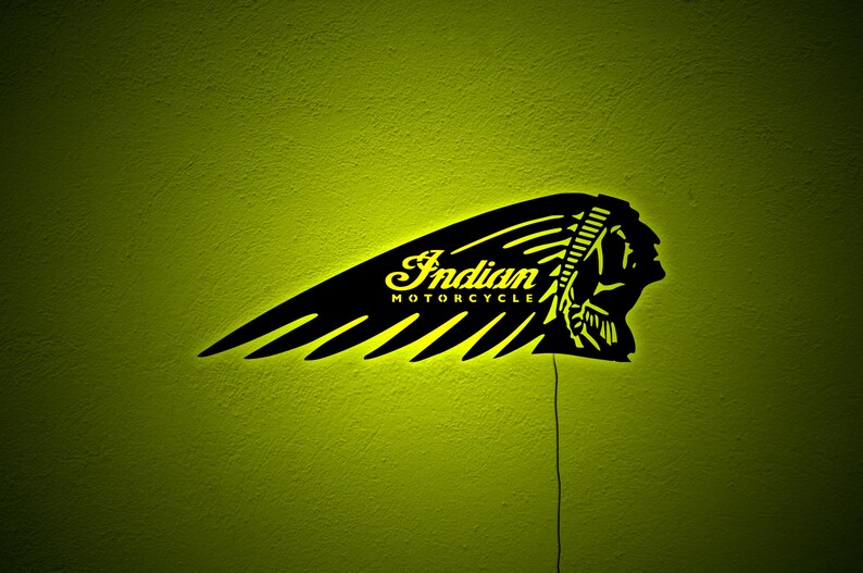 May include: Black Indian Motorcycle logo sign with a yellow glow against a textured green wall. The sign features the brand name and a Native American profile within a wing design.