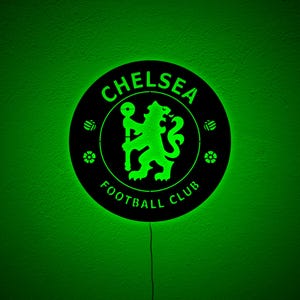 Puede incluir: Un letrero redondo negro del Chelsea Football Club con un logotipo y texto iluminados en verde. El letrero presenta el emblema del club, un le&oacute;n sosteniendo un bast&oacute;n, y las palabras "Chelsea Football Club". El fondo es verde liso.