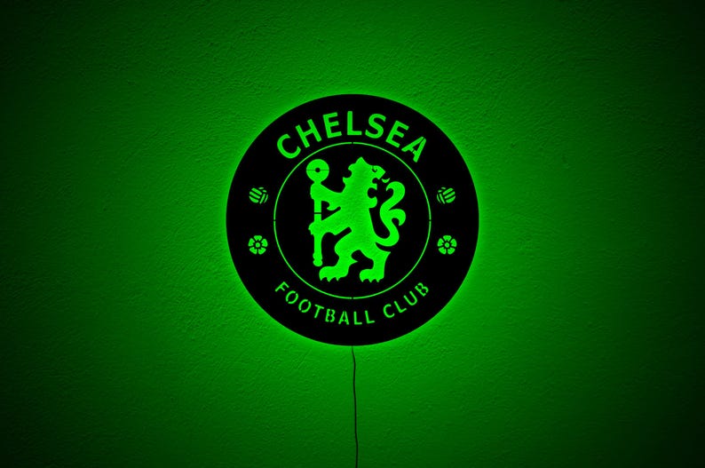 Puede incluir: Un letrero redondo negro del Chelsea Football Club con un logotipo y texto iluminados en verde. El letrero presenta el emblema del club, un le&oacute;n sosteniendo un bast&oacute;n, y las palabras "Chelsea Football Club". El fondo es verde liso.