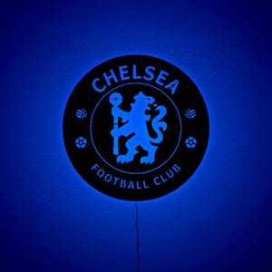 Puede incluir: Un letrero redondo negro del Chelsea Football Club con iluminaci&oacute;n LED azul. El escudo del club, con un le&oacute;n sosteniendo un bast&oacute;n, est&aacute; iluminado en azul. El letrero est&aacute; montado en una pared azul oscuro.