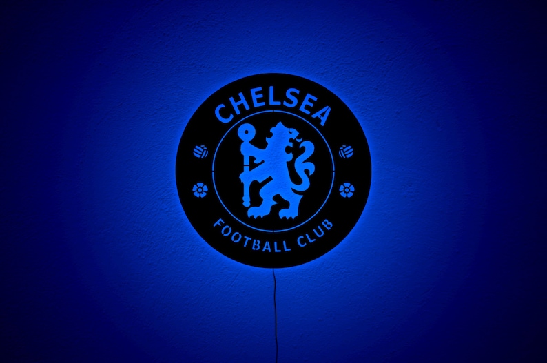 Puede incluir: Un letrero redondo negro del Chelsea Football Club con iluminaci&oacute;n LED azul. El escudo del club, con un le&oacute;n sosteniendo un bast&oacute;n, est&aacute; iluminado en azul. El letrero est&aacute; montado en una pared azul oscuro.
