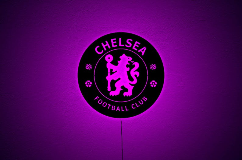 Puede incluir: Un letrero redondo negro del Chelsea Football Club con un resplandor p&uacute;rpura. El nombre y el emblema del club est&aacute;n en el centro, con un le&oacute;n p&uacute;rpura. El letrero est&aacute; iluminado con una luz p&uacute;rpura.