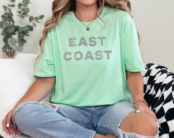 May include: A pale green t-shirt with the text "EAST COAST" in grey. The shirt is paired with ripped blue jeans. The image depicts casual clothing.