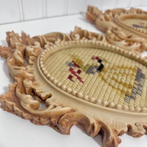 K&ouml;nnte beinhalten: Zwei ovale Vintage-Wandplatten mit verzierten, cremefarbenen Rahmen. Jede Platte zeigt ein Gobelin-Design mit einem Vogelmotiv in Gelb-, Rot-, Schwarz- und Graut&ouml;nen. Die Rahmen haben ein detailliertes Rankenmuster.