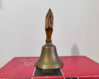 May include: A vintage brass bell with a wooden handle. The handle features a dark brown base with a lighter, textured wood section. The bell sits on a red book with gold lettering. The bell is a classic decorative item.