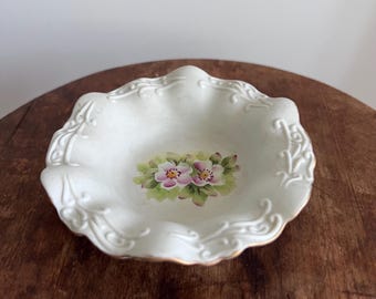 May include: A small, white, scalloped-edge dish with a floral design. The dish features a hand-painted image of pink and white flowers with green leaves in the center. The rim has a gold trim and decorative scrollwork.