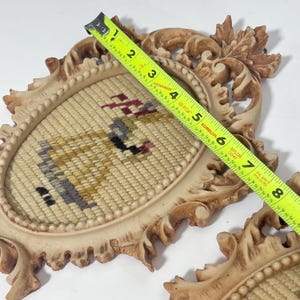 K&ouml;nnte beinhalten: Zwei ovale Vintage-Wandplaketten mit Nadelspitzeins&auml;tzen. Die Plaketten haben verzierte, cremefarbene Rahmen. Die Nadelspitze hat einen beigefarbenen Hintergrund mit einem farbenfrohen Design. Die Plaketten sind etwa 15 cm breit.