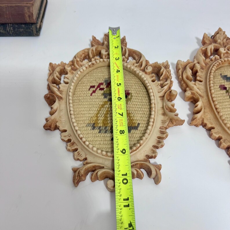 K&ouml;nnte beinhalten: Zwei gerahmte Vintage-Nadelmalereien. Die verzierten cremefarbenen Rahmen sind etwa 25 cm hoch. Die Nadelmalerei zeigt eine Figur in einem gelben Kleid mit roten Akzenten. Der Hintergrund ist hellbeige.