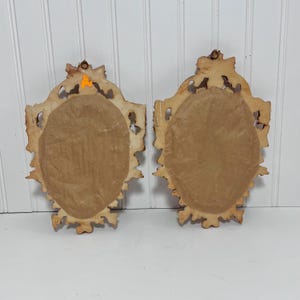 K&ouml;nnte beinhalten: Zwei ovale, verzierte Vintage-Wandspiegel mit hellbraunem, abgenutztem Finish. Die R&uuml;ckseiten sind mit braunem Papier bedeckt. Die Rahmen haben ein detailliertes, geschnitztes Design. Die Spiegel werden vor einem wei&szlig;en Hintergrund mit vertikalen Lamellen gezeigt.