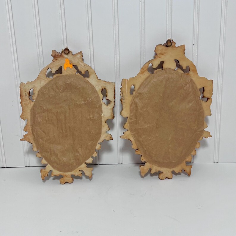 K&ouml;nnte beinhalten: Zwei ovale, verzierte Vintage-Wandspiegel mit hellbraunem, abgenutztem Finish. Die R&uuml;ckseiten sind mit braunem Papier bedeckt. Die Rahmen haben ein detailliertes, geschnitztes Design. Die Spiegel werden vor einem wei&szlig;en Hintergrund mit vertikalen Lamellen gezeigt.