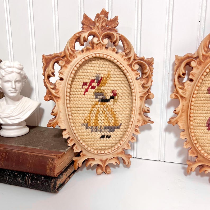K&ouml;nnte beinhalten: Zwei ovale Vintage-Wandbeh&auml;nge mit verzierten, hellbraunen Rahmen. Jedes zeigt ein Gobelin-Design einer Frau in einem gelben Kleid, die einen rot-wei&szlig;en Gegenstand h&auml;lt. Die Rahmen haben ein detailliertes, geschnitztes Design.