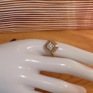 Può includere: Un anello dorato con un elemento centrale a forma di diamante quadrato. Il centro presenta un gruppo di piccole pietre trasparenti. L'anello è mostrato su una mano bianca, con un tessuto a righe marrone e bianco sullo sfondo.