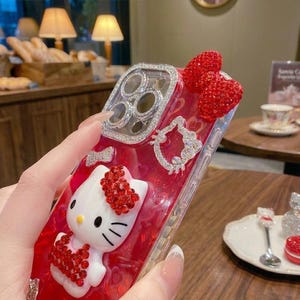 May include: A red and clear phone case featuring a white Hello Kitty figure with red rhinestone accents. The case has a rhinestone-encrusted frame around the camera and two red rhinestone hearts. A rhinestone Hello Kitty outline is also present.