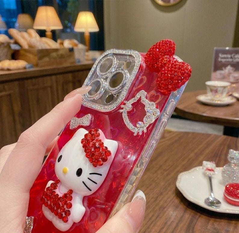May include: A red and clear phone case featuring a white Hello Kitty figure with red rhinestone accents. The case has a rhinestone-encrusted frame around the camera and two red rhinestone hearts. A rhinestone Hello Kitty outline is also present.