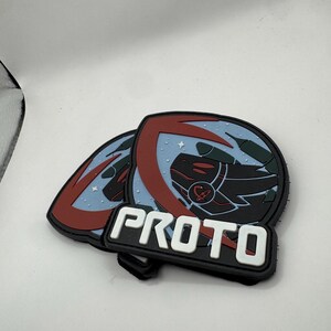 May include: Two black and white PROTO patches. Each patch has a circular design with a red and blue border, a black and red graphic, and the word "PROTO" in white block letters.