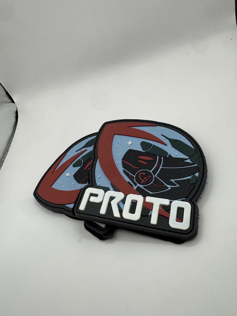 May include: Two black and white PROTO patches. Each patch has a circular design with a red and blue border, a black and red graphic, and the word "PROTO" in white block letters.