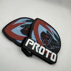 May include: A set of three black, red, and blue PVC patches. Two patches feature a stylized dragon design with a red arc and a light blue background. A rectangular patch below reads "PROTO" in white block letters.