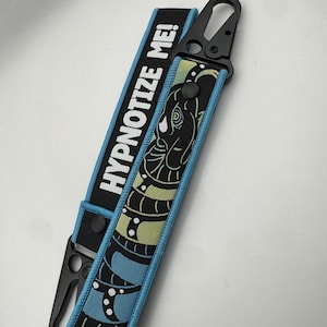 May include: A black and blue key fob with the text "HYPNOTIZE ME!". The fob features a snake design in green and blue, with a black clasp. The edges are trimmed in light blue.