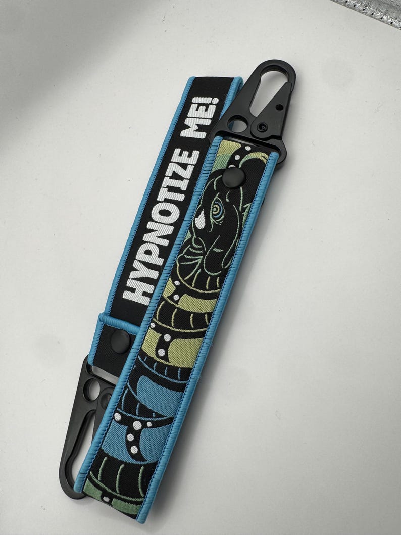 May include: A black and blue key fob with the text "HYPNOTIZE ME!". The fob features a snake design in green and blue, with a black clasp. The edges are trimmed in light blue.