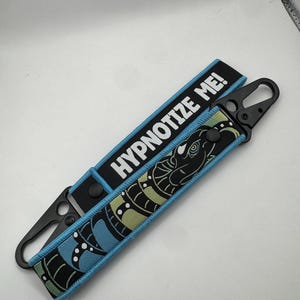 May include: A black and blue keychain strap with the text "HYPNOTIZE ME!". The strap features a stylized snake design in black, blue, and green. It has a black metal clasp for attachment.