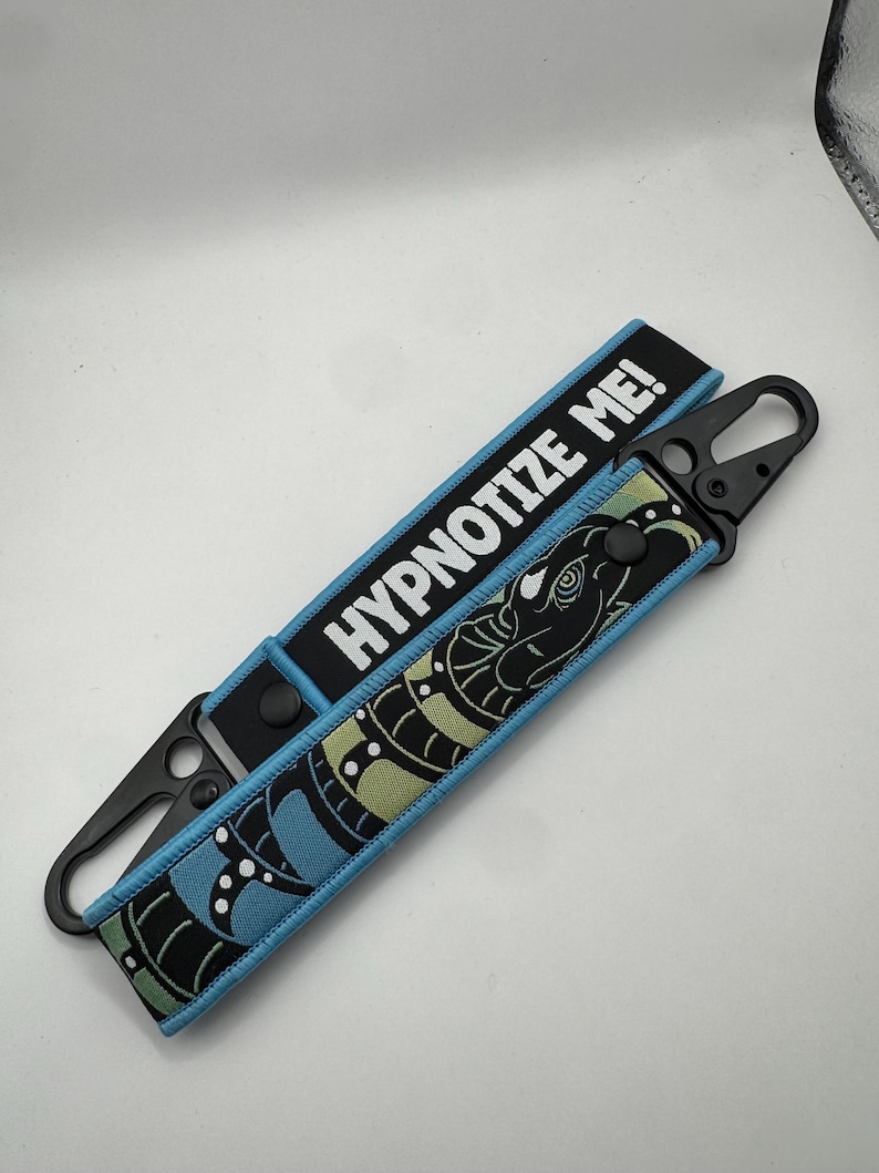 May include: A black and blue keychain strap with the text "HYPNOTIZE ME!". The strap features a stylized snake design in black, blue, and green. It has a black metal clasp for attachment.