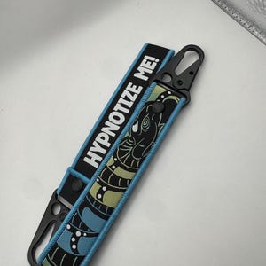 May include: A black and blue keychain wrist strap with the text "HYPNOTIZE ME!" in white. The strap features a black and green snake design. The strap has a black metal clasp.