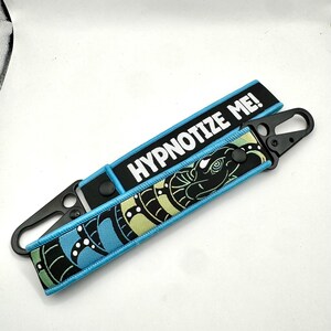 May include: Two black keychains with a blue and black design. One keychain has the text "HYPNOTIZE ME!" in white. The other keychain has a blue, green, and black snake design.